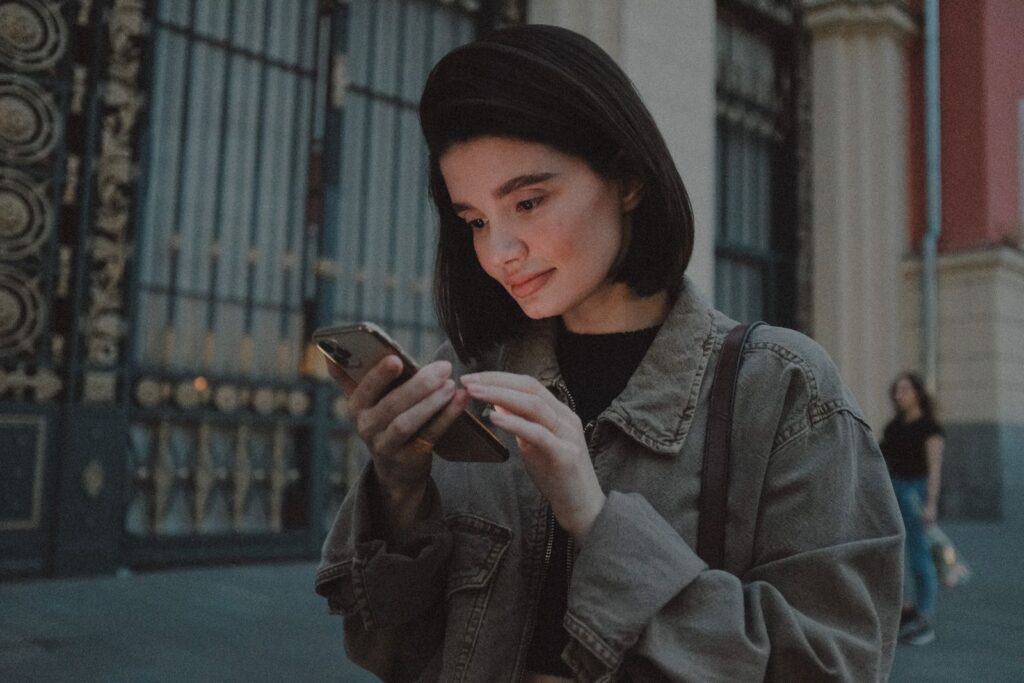 Young female in denim jacket browsing smartphone while standing on historic street in evening