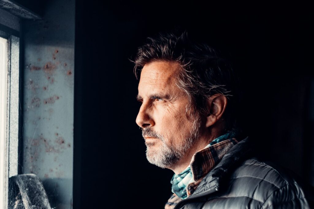 An elderly man with facial hair gazes pensively out a window in a dimly lit room.