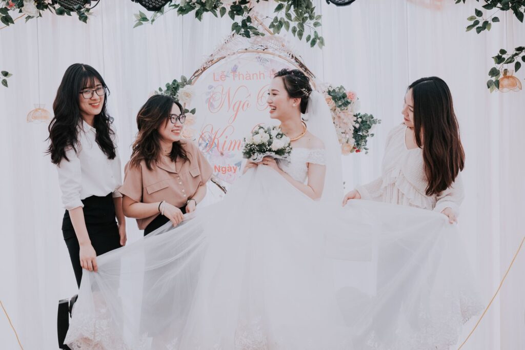 Laughing young Asian woman in bridal dress with bouquet and girlfriends against flowers in tent