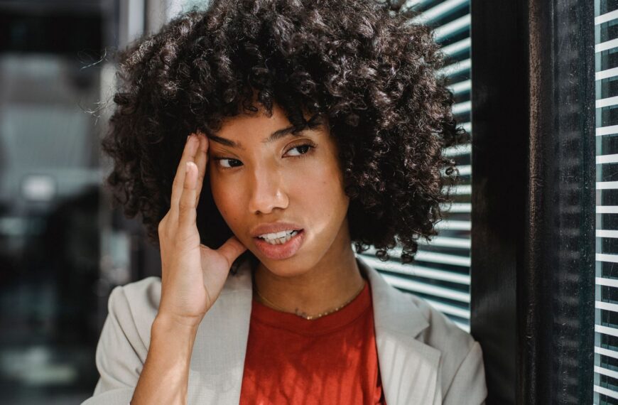 A businesswoman with curly hair expresses stress by her office window, touching her forehead.