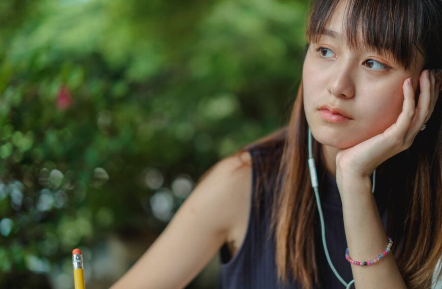 Pensive Asian female student listening to music via earphones while doing homework and taking notes with pencil
