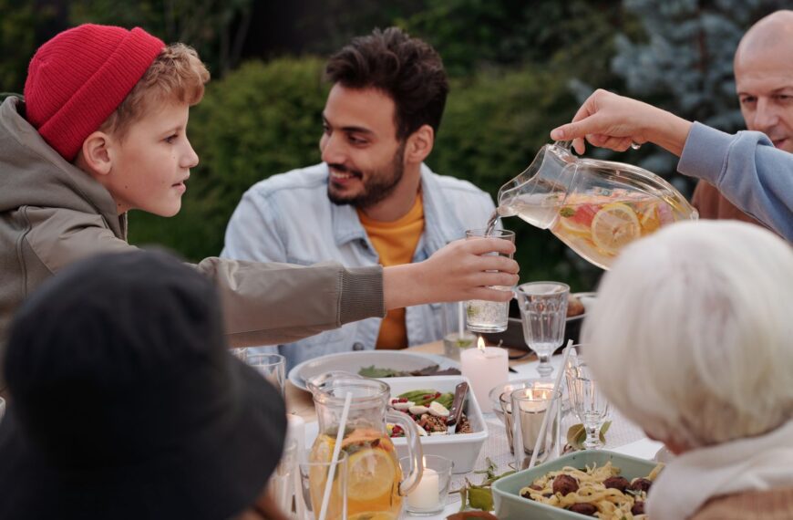 Faceless female pouring fruit beverage into glass of boy in warm cloth while cheerful multiethnic family having dinner outdoors