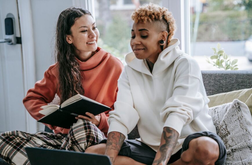 Positive Hispanic female and smiling African American female sitting on couch and using laptop and notebook