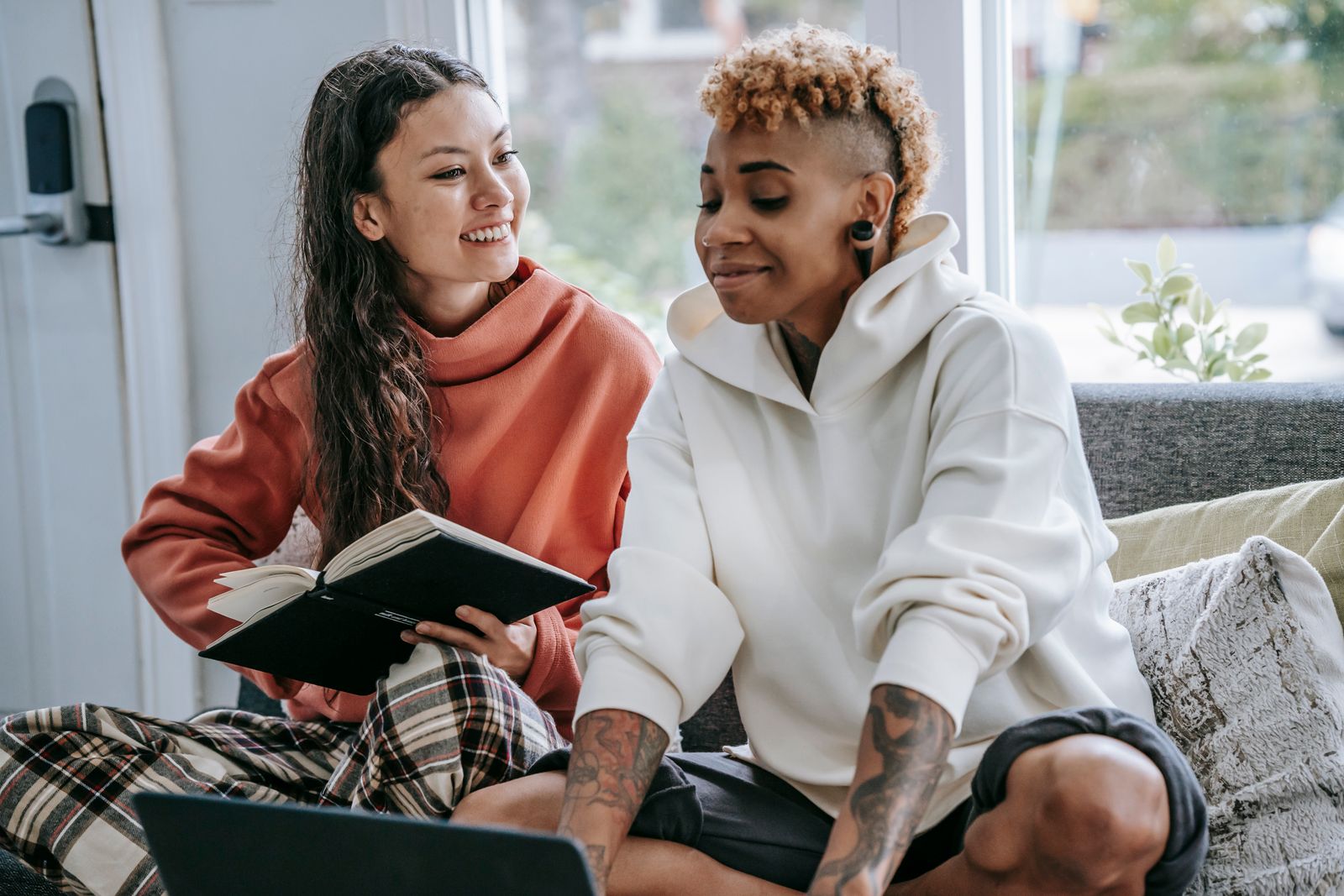 Positive Hispanic female and smiling African American female sitting on couch and using laptop and notebook