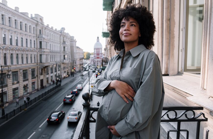 A pregnant woman stands on a balcony overlooking a bustling city street, radiating tranquility.