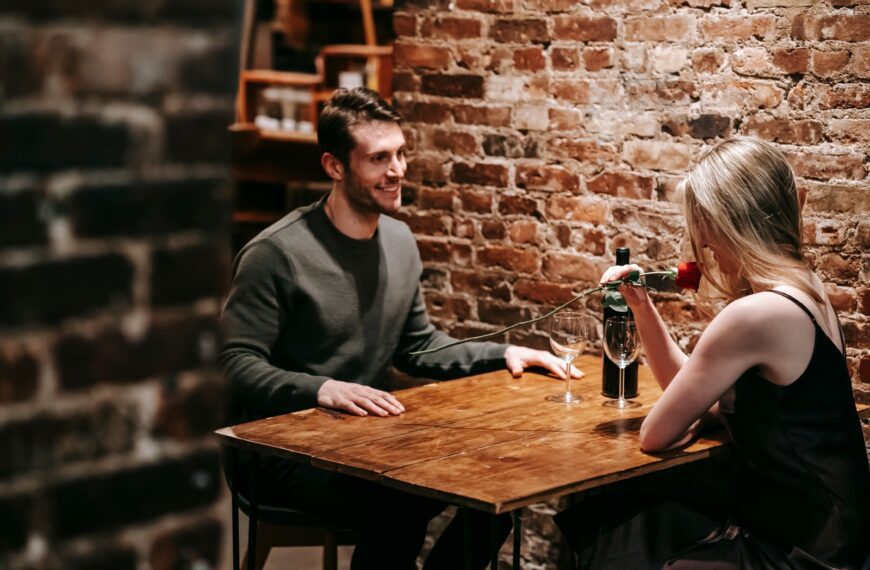 Young couple in elegant clothes sitting in light cafeteria at wooden table near brick wall and having romantic dinner with red rose and wine near glasses