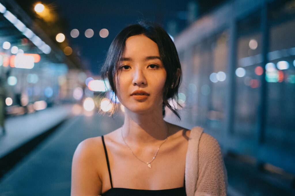 Slim ethnic female looking at camera on blurred background of urban illumination on street