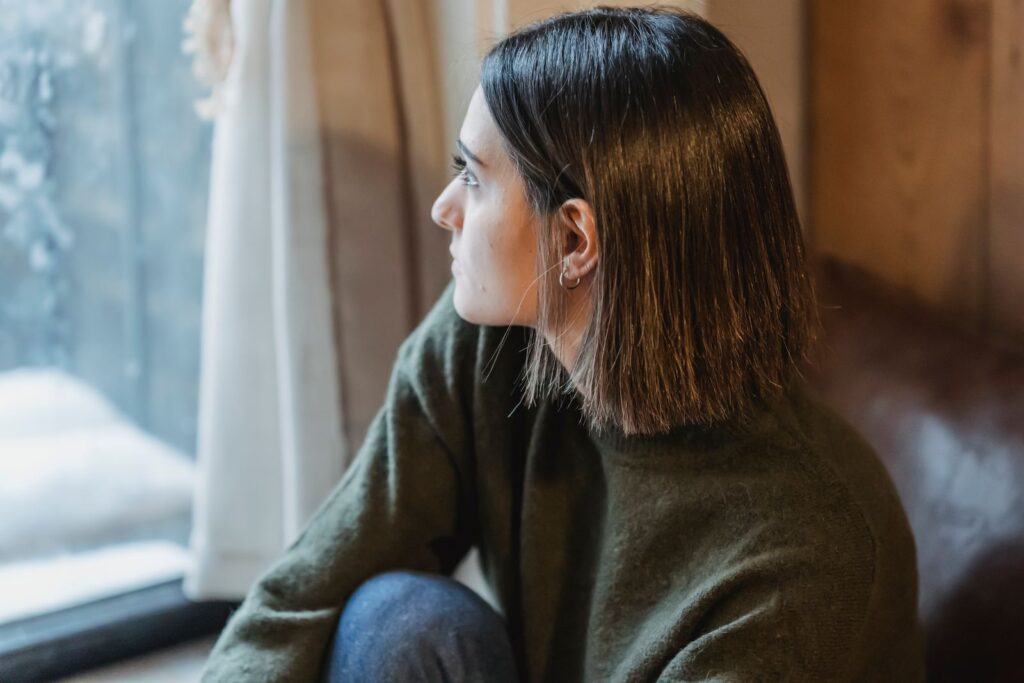 From above of dreamy female in casual wear sitting on windowsill and thoughtfully looking away