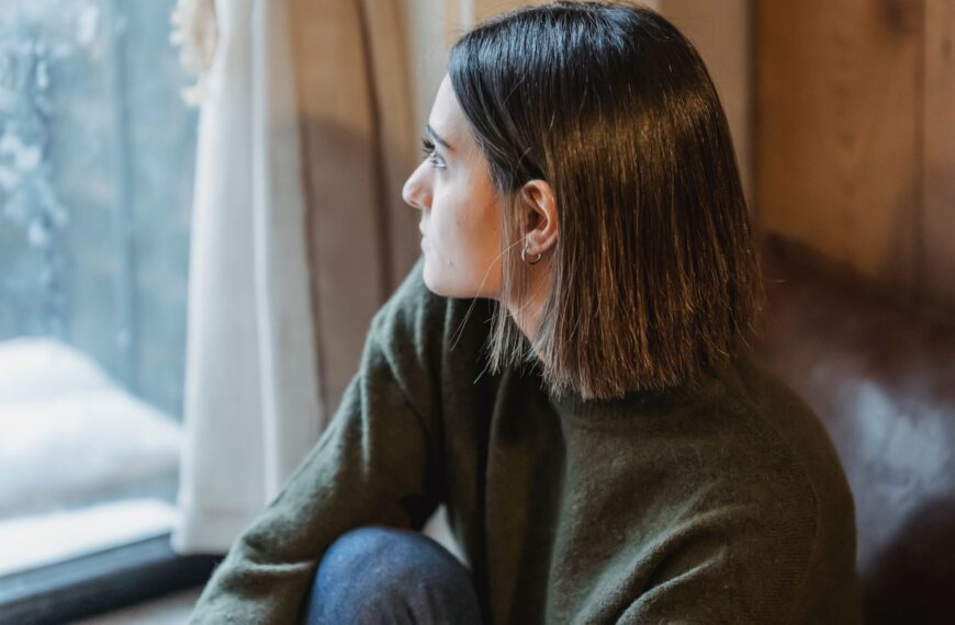 From above of dreamy female in casual wear sitting on windowsill and thoughtfully looking away