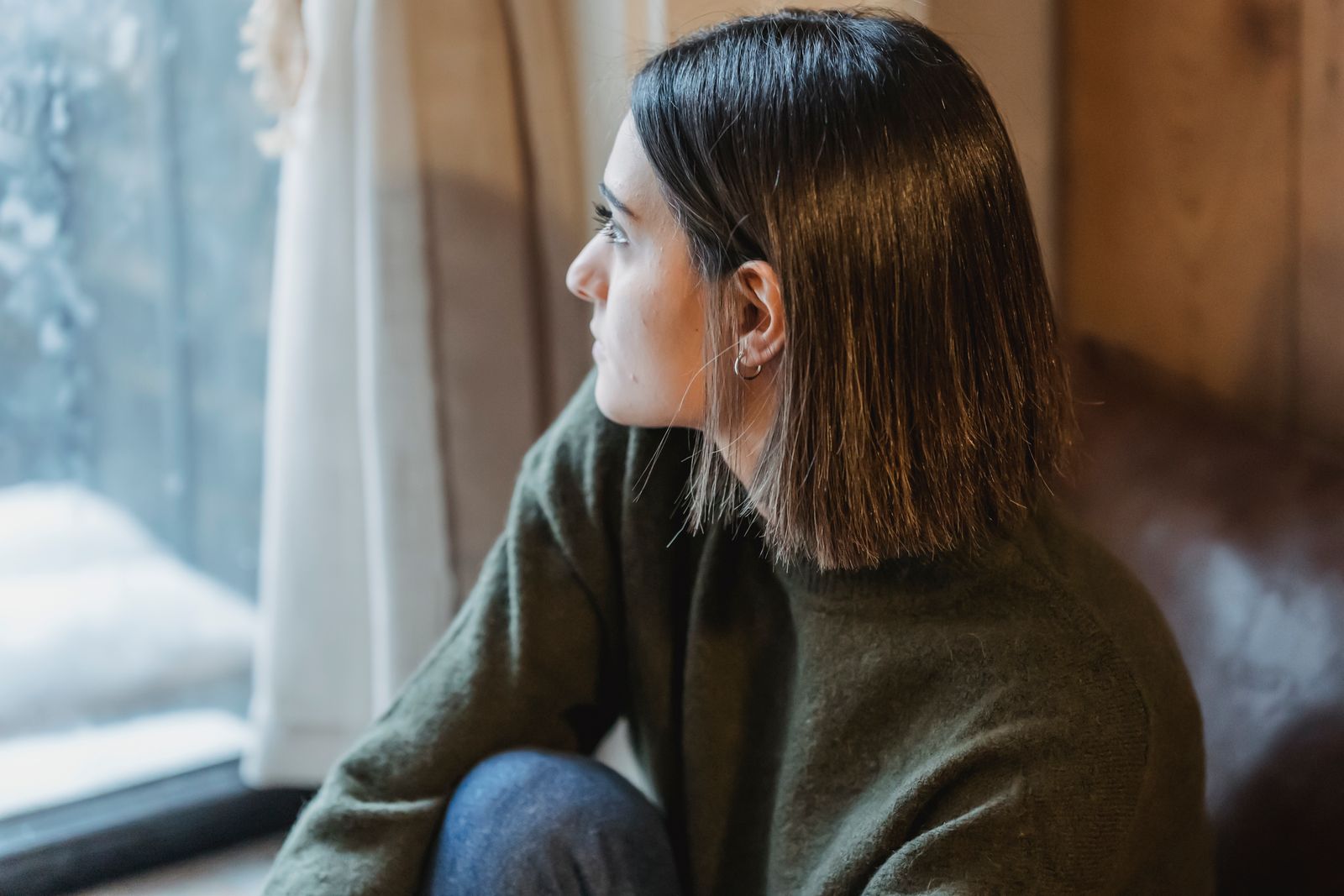 From above of dreamy female in casual wear sitting on windowsill and thoughtfully looking away