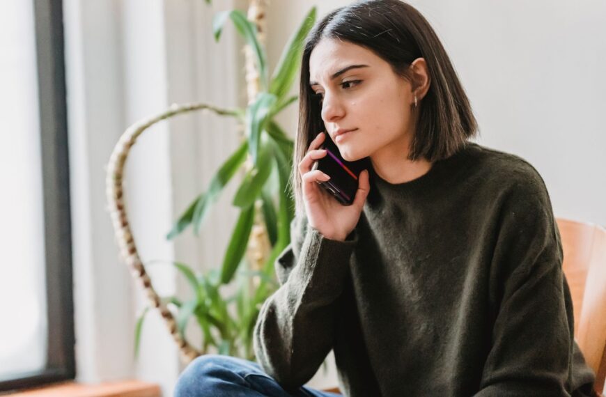 Calm young ethnic female millennial with dark hair in casual clothes having phone conversation while sitting on comfortable armchair at home in daylight