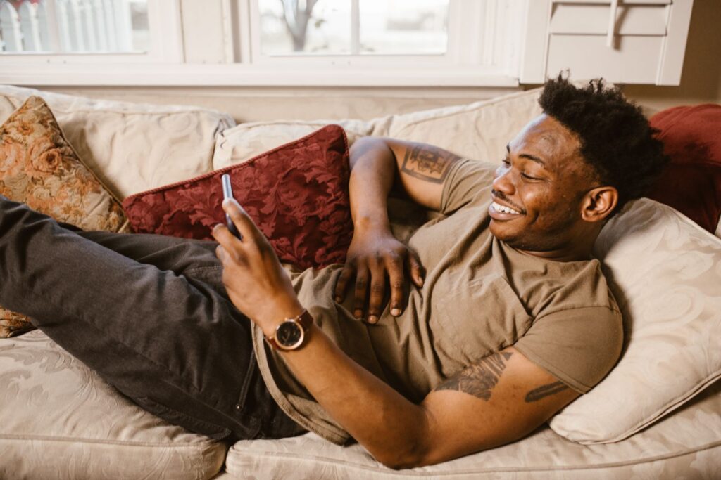 Smiling man lying on a couch using smartphone, enjoying relaxation at home.
