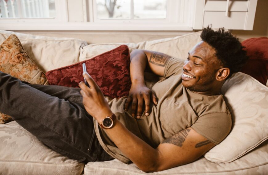 Smiling man lying on a couch using smartphone, enjoying relaxation at home.