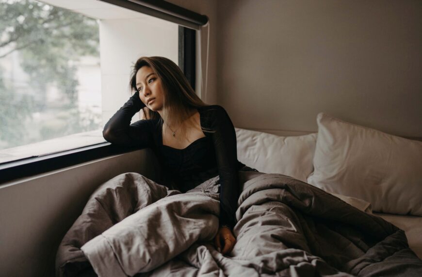 A young woman in a cozy room, pondering by the window, wrapped in blankets.