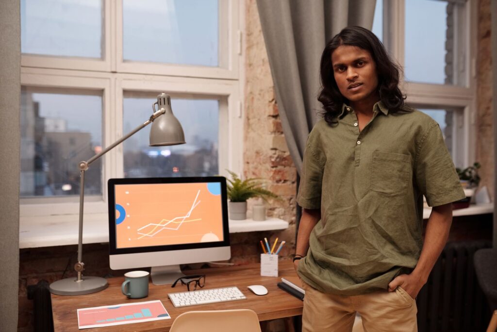 Asian man stands in a modern office with a computer and desk, showcasing a professional work environment.