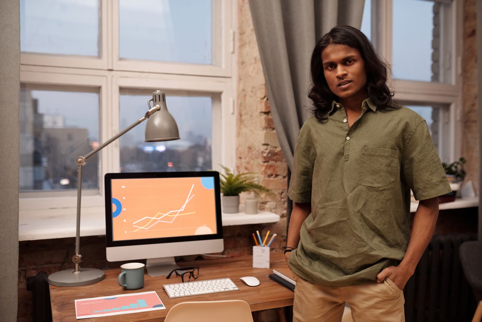 Asian man stands in a modern office with a computer and desk, showcasing a professional work environment.