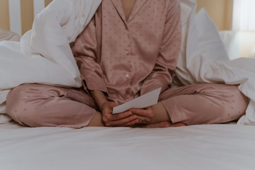 A person in pajamas sits on a bed, enveloped in blankets while holding a letter.