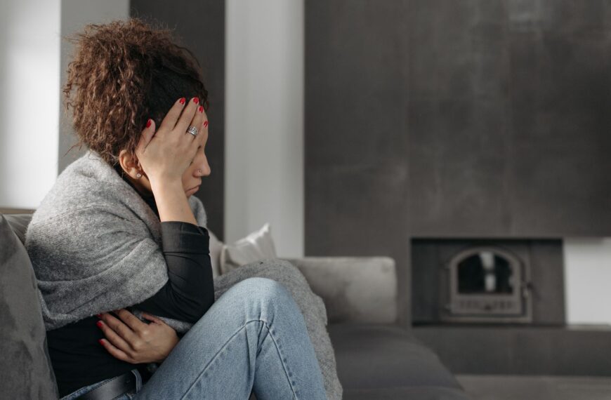 A woman sits on a couch indoors, wrapped in a blanket, holding her head in discomfort.