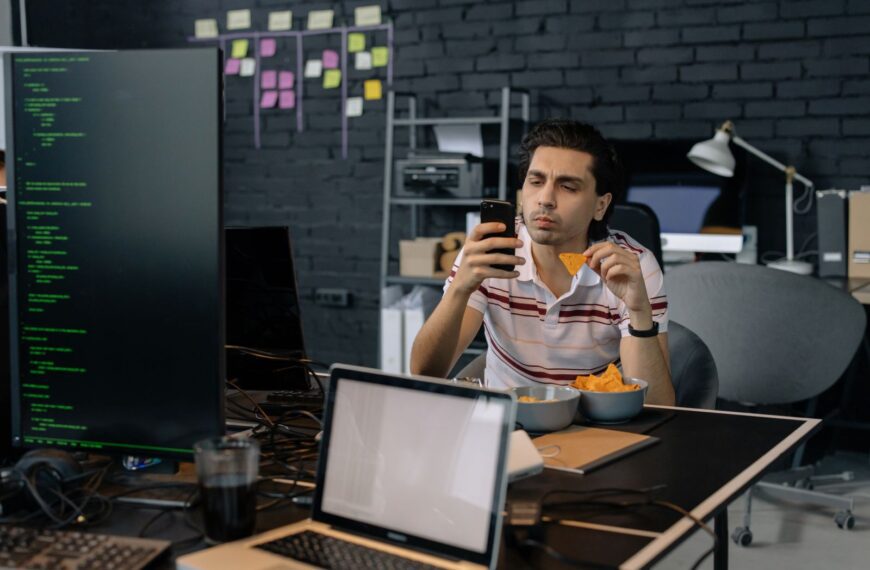 A programmer checks his smartphone while eating snacks at a modern tech office desk.