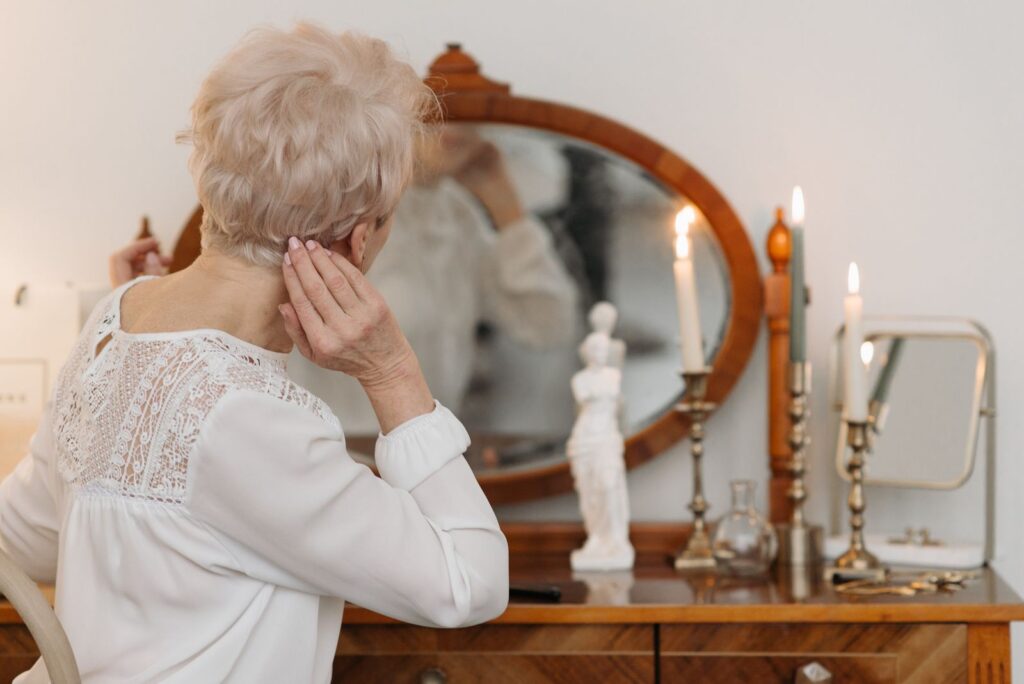 Senior woman with short hair styles herself at a vanity table with candles.