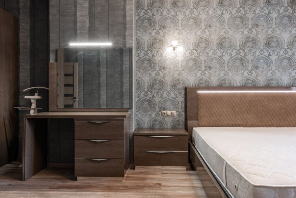 Comfortable bed with white mattress placed at wall with glowing sconce wallpaper and wooden cupboard with mirror in stylish bedroom