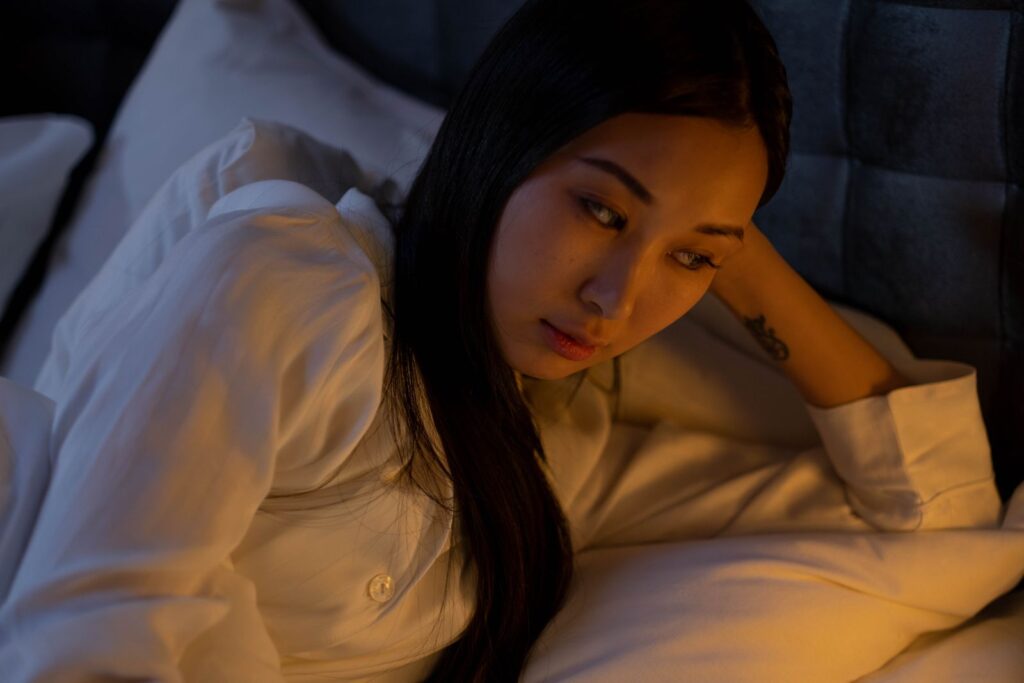 Asian woman in a serene bedroom setting, deep in thought, offering a sense of tranquility.