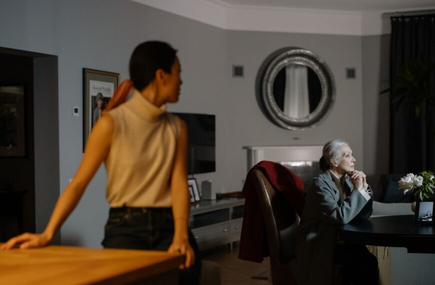 Senior woman deep in thought at a table with a younger woman in focus background, evoking a quiet, introspective mood.