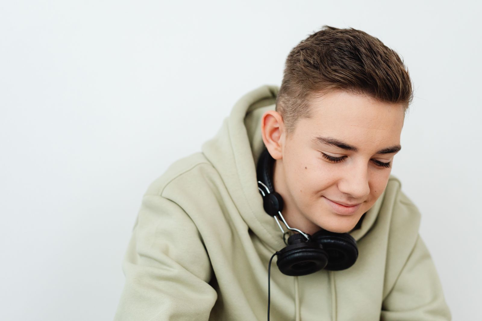 A young man smiling and looking down, wearing a hoodie and headphones against a white background.
