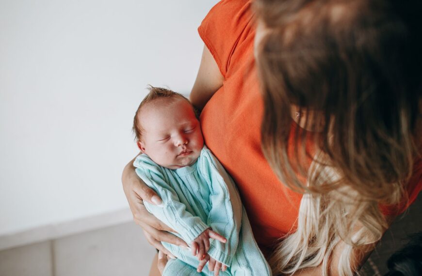 Crop anonymous mother with long hair with sleeping baby in arms sitting in light room near white wall at home