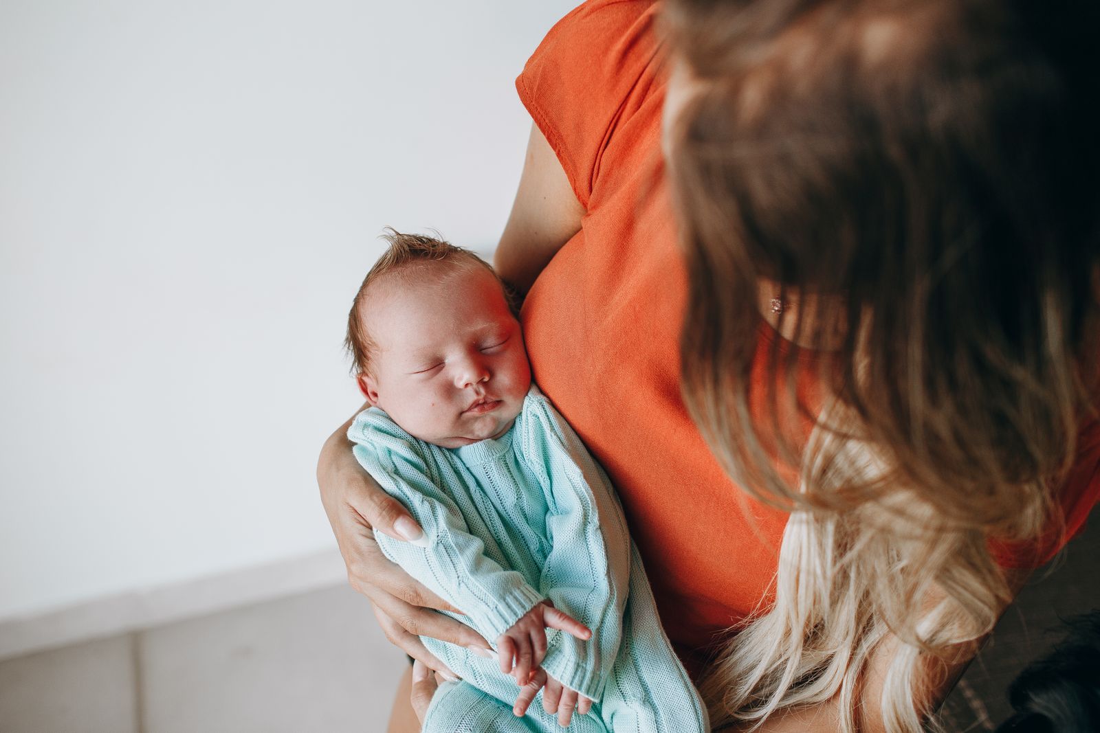 Crop anonymous mother with long hair with sleeping baby in arms sitting in light room near white wall at home