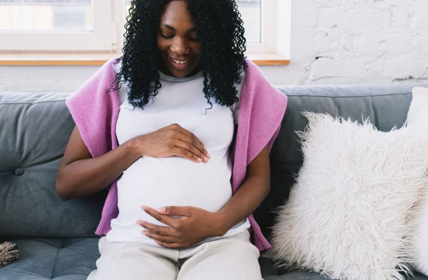 Cheerful pregnant African American female touching tummy gently while sitting on comfortable sofa with pillows in light room at home