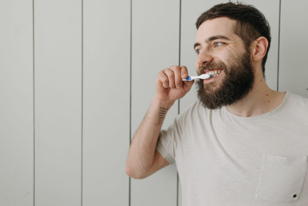Adult man with a beard brushing his teeth indoors with a manual toothbrush.