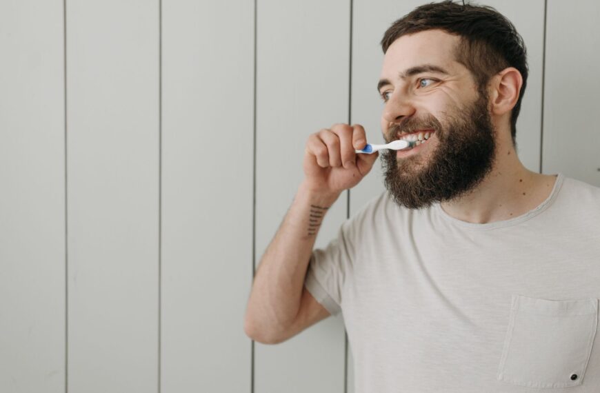 Adult man with a beard brushing his teeth indoors with a manual toothbrush.