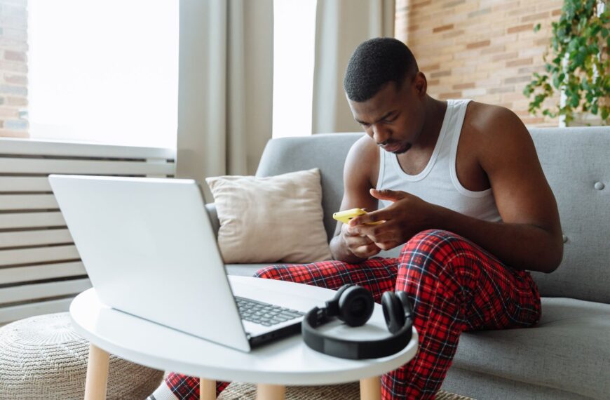 Man sitting on sofa, multitasking with laptop and smartphone in cozy living room.