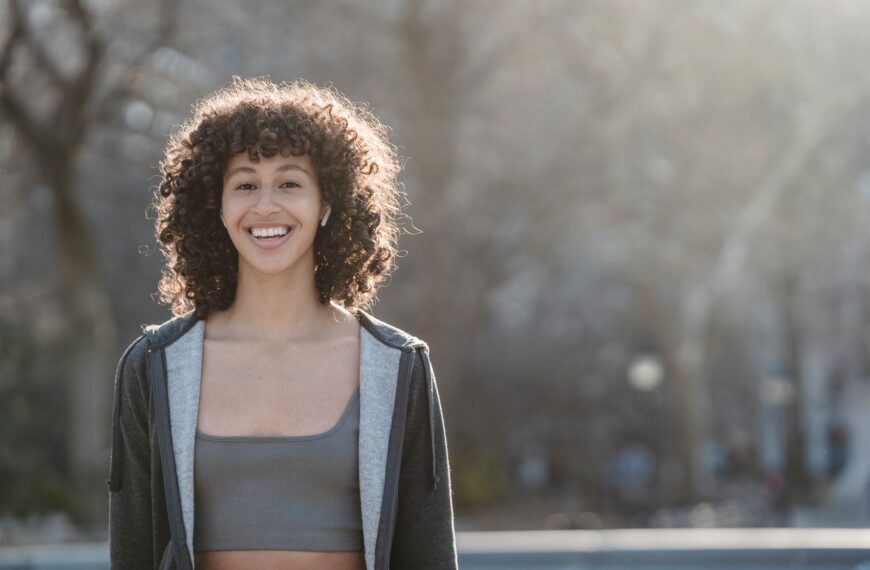Delighted ethnic sportswoman in activewear in wireless earbuds looking at camera while standing on street against blurred background during training
