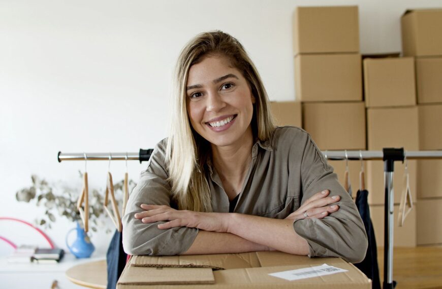 Happy woman managing her online store with packages and clothing rack.