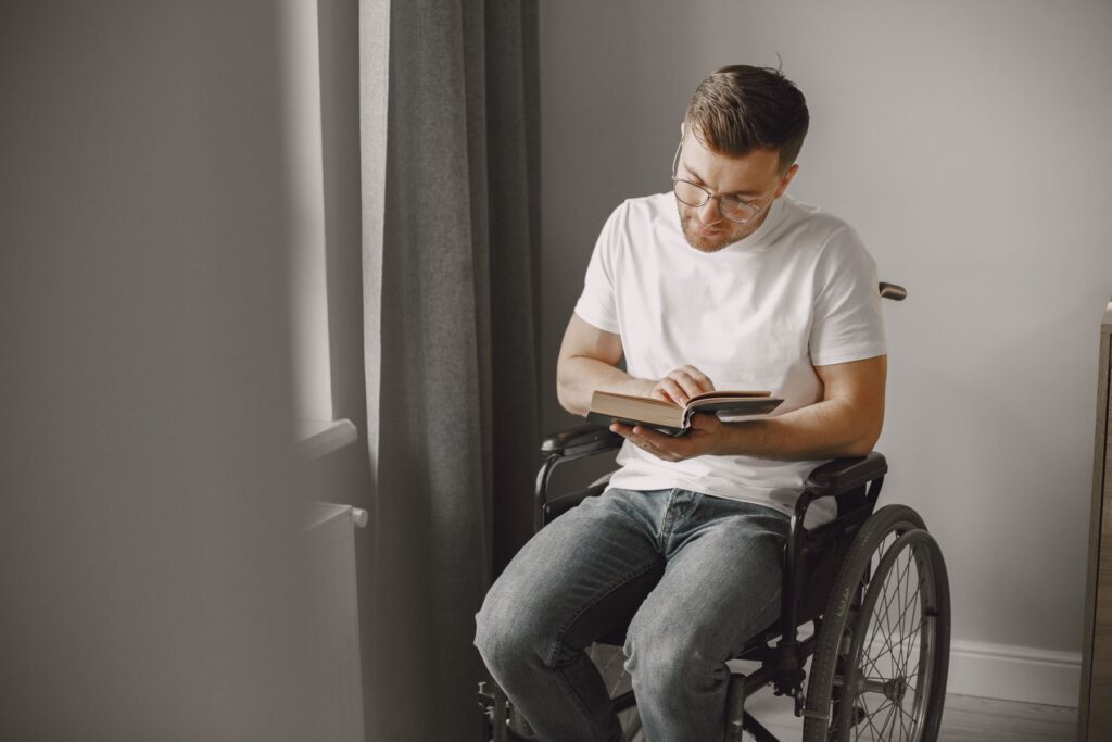 A man in a wheelchair reads a book by a window in an indoor setting.