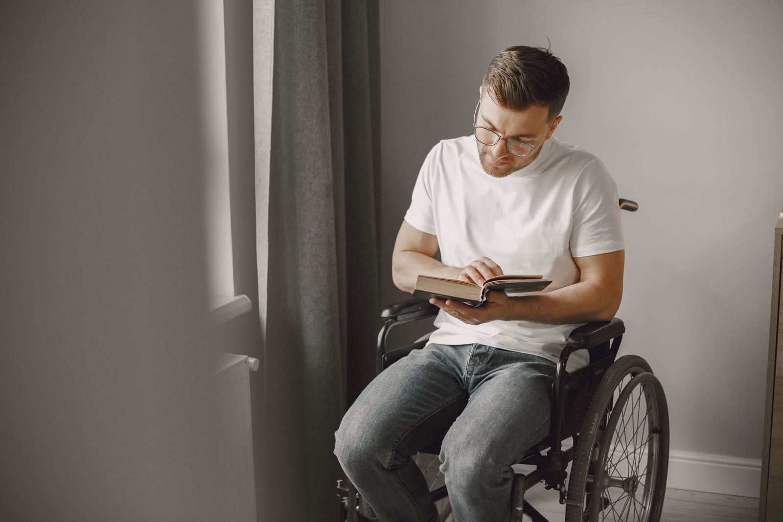 A man in a wheelchair reads a book by a window in an indoor setting.