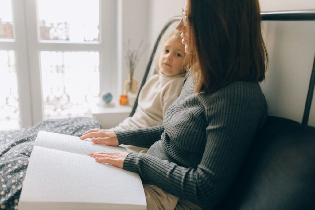 A woman and child reading a braille book in a cozy bedroom setting, enhancing child development.
