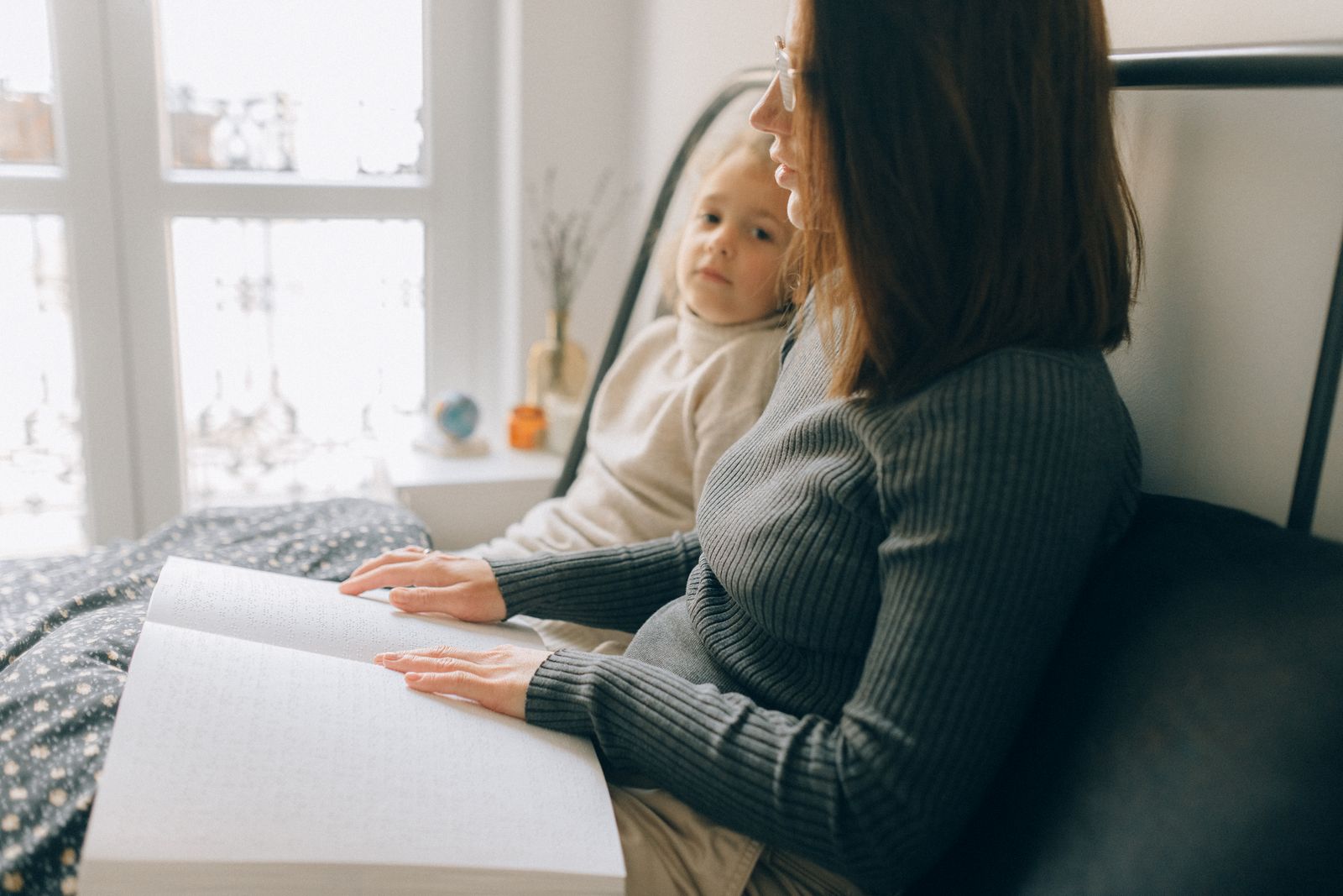 A woman and child reading a braille book in a cozy bedroom setting, enhancing child development.