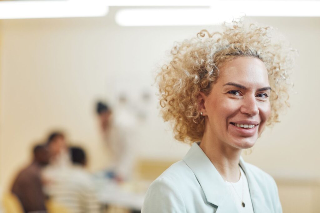 Close-up of a smiling woman with curly blonde hair in an office environment.
