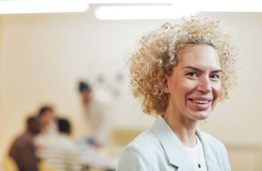 Close-up of a smiling woman with curly blonde hair in an office environment.
