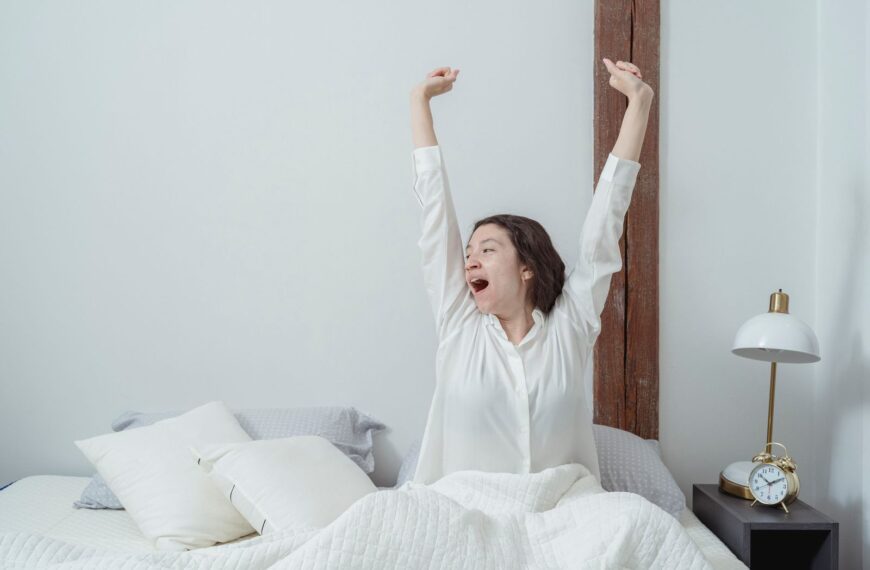Sleepy brunette sitting on white comfortable bed under blanket and stretching arms up while yawning and looking away during morning time at home