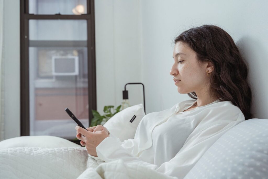 Side view of young content female with long dark hair in white nightwear sitting on white bed and browsing mobile phone in morning time