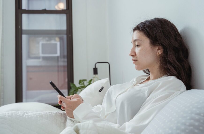 Side view of young content female with long dark hair in white nightwear sitting on white bed and browsing mobile phone in morning time