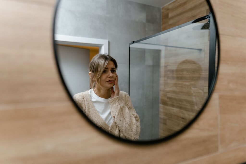 Woman in neutral sweater looking at herself in a bathroom mirror, creating a serene and introspective mood.