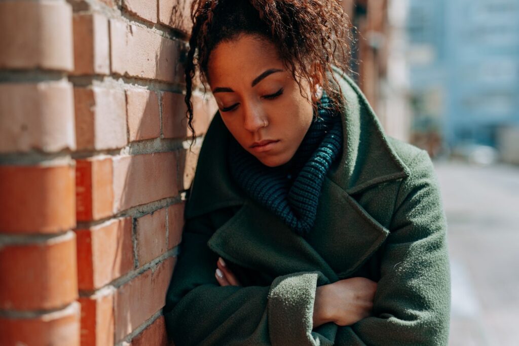 Young woman in green coat leaning on brick wall, looking thoughtful.