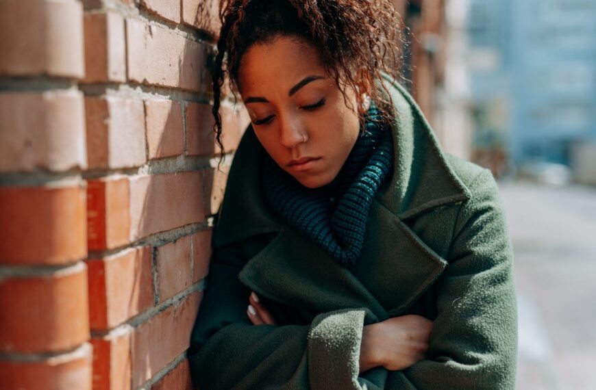 Young woman in green coat leaning on brick wall, looking thoughtful.
