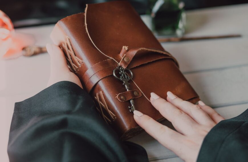 A close-up of a hand holding a leather-bound spell book with a key, hinting at a magical theme.
