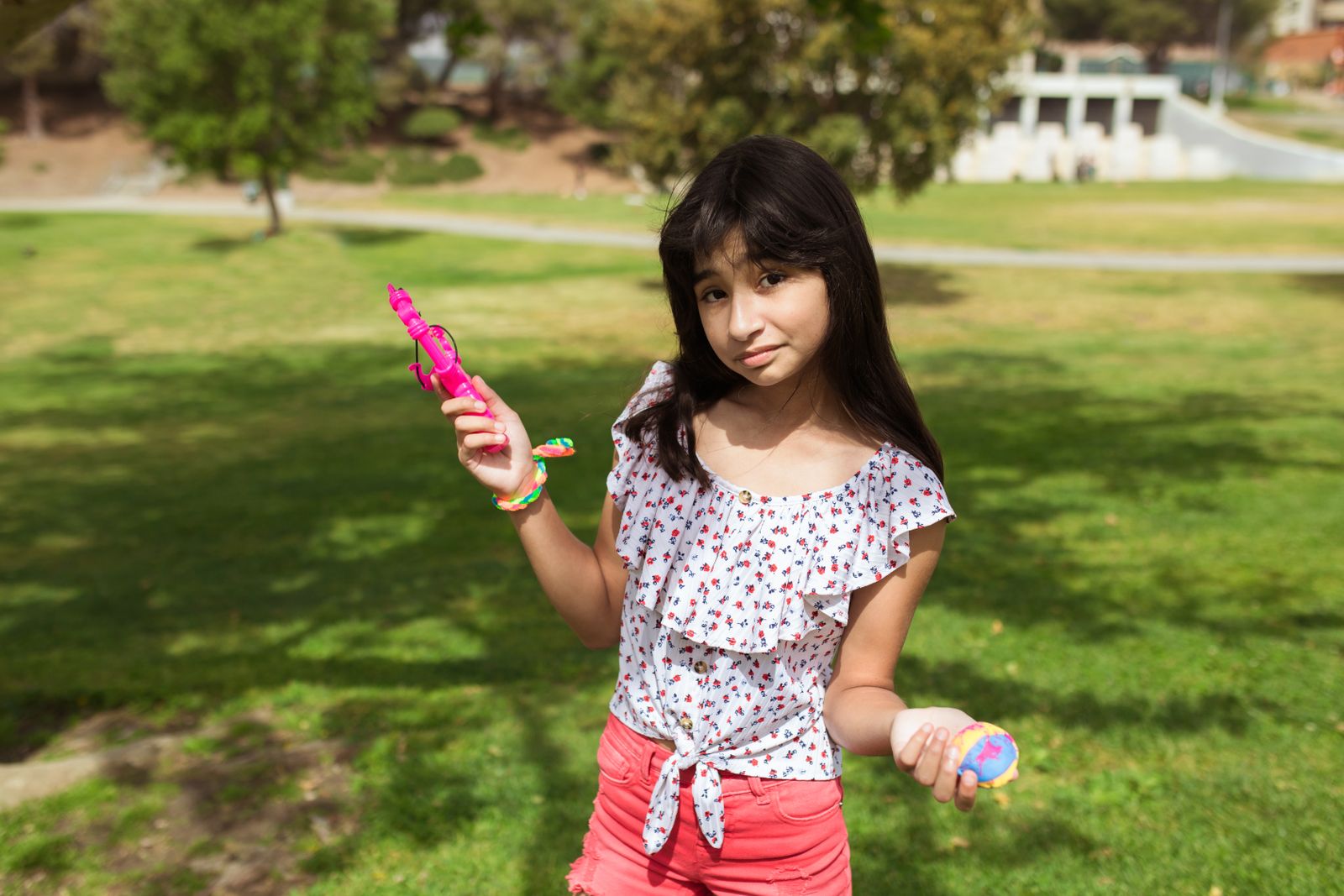 A young girl poses in a sunny park holding colorful plastic toys.